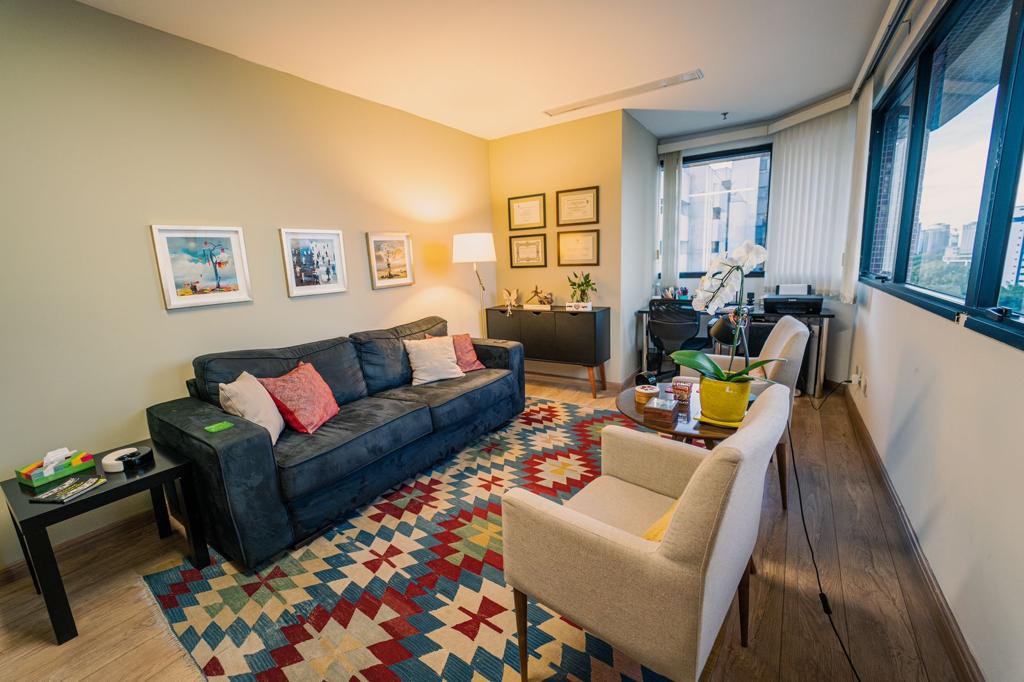 Sala de atendimento com sofá, poltronas claras, tapete colorido e janelas amplas com vista para a cidade
